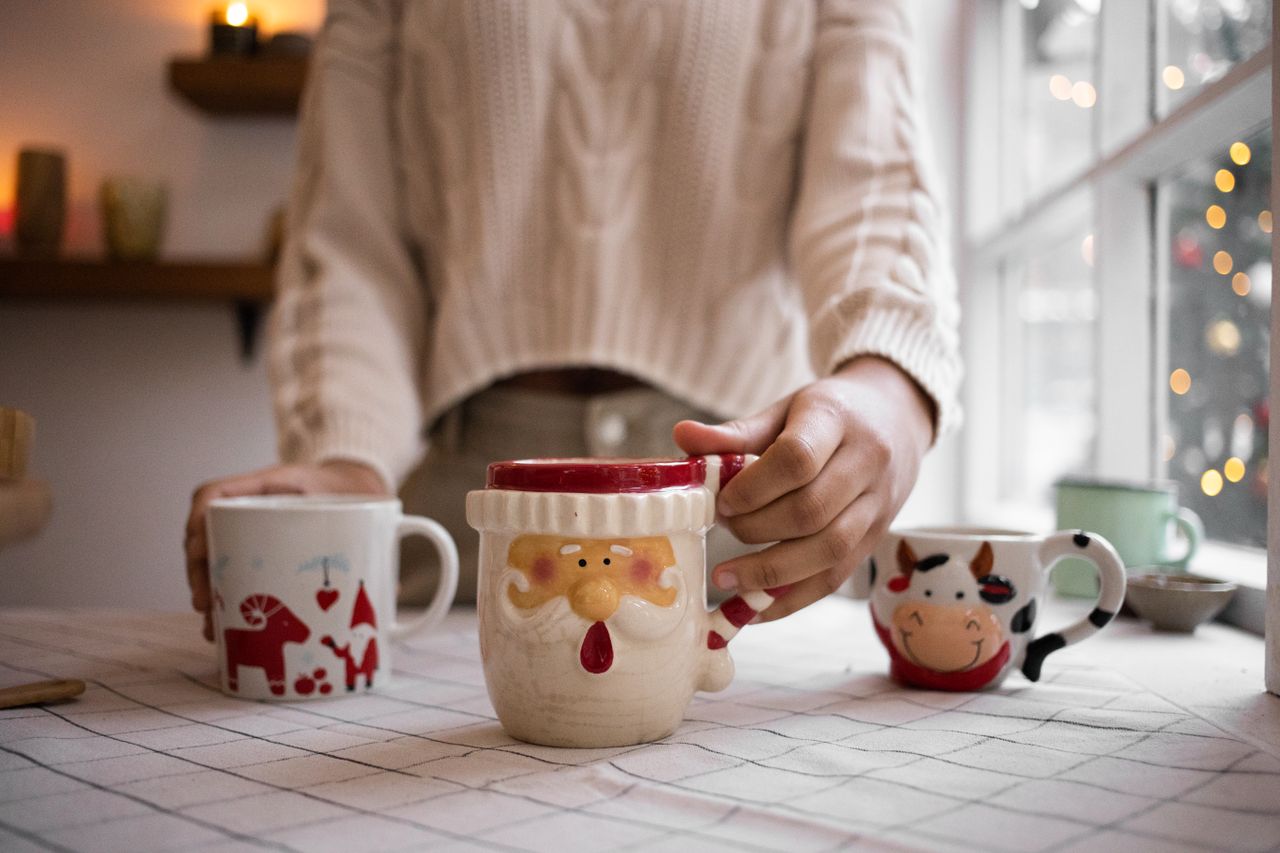 our-story Close-up of hands holding decorative Christmas mugs indoors, capturing the warmth of the holiday season.